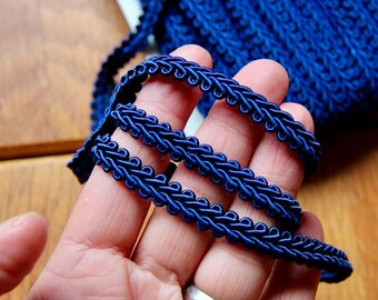 Könnte beinhalten: Nahaufnahme eines dunkelblauen geflochtenen Besatzes mit einem dekorativen Rand. Der Besatz wird in einer Hand gehalten, wodurch die Textur und die Details der Flechtung sichtbar werden.