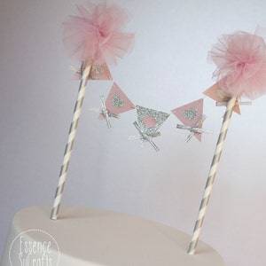 May include: A pink and silver cake topper with a banner of pink triangles with silver glitter hearts. The banner is attached to two striped straws with pink tulle pom-poms on top. The cake topper says "Essence Sucrafts" on the bottom.