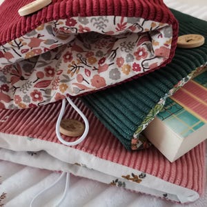 Peut inclure: Trois housses de livre en velours côtelé bordeaux, vert foncé et rose poudré. Chaque housse dispose d'une fermeture élastique blanche avec un bouton en bois rond et une doublure en tissu à motifs floraux dans des tons rose, vert et marron.