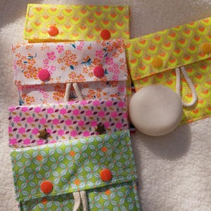 Peut inclure: Plusieurs pochettes en tissu aux motifs colorés et fermetures à bouton-pression. Les pochettes présentent des motifs floraux et géométriques dans les tons rose, vert et jaune. Un savon rond et blanc est posé à côté d'une des pochettes.