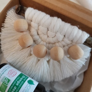 Peut inclure: Une suspension murale en macramé blanche avec des perles en bois et une petite bouteille d'huile essentielle d'orange. L'étiquette de la bouteille indique "voshuiles" et "Orange douce huile essentielle bio".