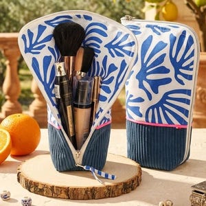 Peut inclure: Deux trousses de toilette verticales avec une partie supérieure en tissu blanc à motif de feuilles bleues abstraites et une base en velours côtelé bleu foncé. Une fine bande rose néon sépare les sections. Une trousse est ouverte, montrant des pinceaux.