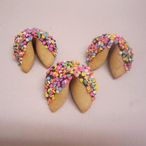May include: Three golden-brown fortune cookies, each partially dipped in dark chocolate and covered in a dense layer of small, round, multi-colored sprinkles in shades of pink, yellow, green, blue, and orange, arranged on a plain surface.