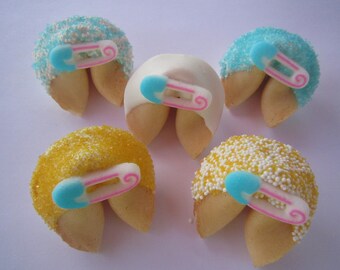 May include: Six fortune cookies decorated with blue and yellow sprinkles and a pink and blue baby safety pin. The cookies are arranged in a semi-circle on a white surface.