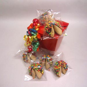 May include: A red box filled with individually wrapped fortune cookies. The cookies are dipped in chocolate and covered in colorful sprinkles. A bundle of colorful ribbons is attached to the box.