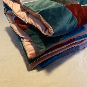 Puede incluir: Un edredón de retazos plegado con cuadrados de varios colores, incluyendo rosa, verde azulado, burdeos y gris. El edredón parece estar hecho de un material suave y aterciopelado, y está apilado ordenadamente sobre una cama.