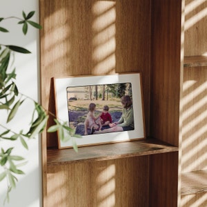 May include: A framed photograph of three people sitting on a grassy area. A woman is sitting on the right, a girl is sitting on the left, and a boy is sitting in the middle. The photo is set on a wooden shelf.