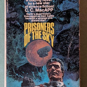 Puede incluir: Un libro de bolsillo de ciencia ficción vintage titulado Prisoners of the Sky de C. C. MacAPP. La portada presenta un fondo cósmico azul oscuro y negro, un gran planeta naranja, una aeronave flotante y una persona con traje blanco y gafas rojas.