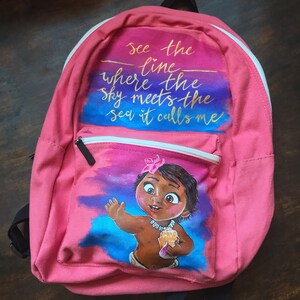 May include: Pink canvas backpack with a hand-painted design featuring a cartoon character from the movie Moana. The design includes a sunset sky with the words "See the line where the sky meets the sea it calls me".