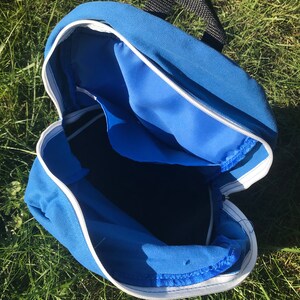 Pu&ograve; includere: Uno zaino in tela blu con una cerniera bianca e un manico nero. Lo zaino &egrave; aperto e vuoto.