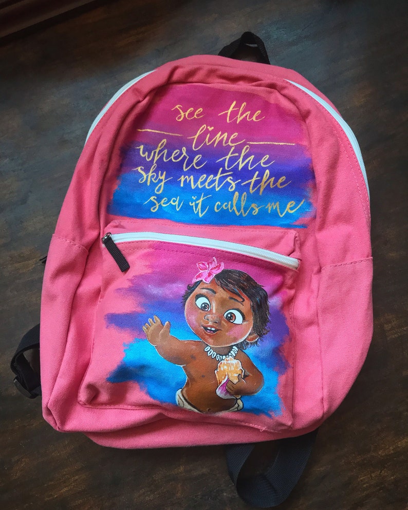 May include: Pink canvas backpack with a hand-painted design featuring a cartoon character from the movie Moana. The design includes a sunset sky with the words "See the line where the sky meets the sea it calls me".