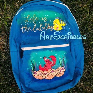 Pu&ograve; includere: Uno zaino in tela blu con un design dipinto a mano con i personaggi Sebastian e Flounder del film Disney La Sirenetta. Lo zaino ha il testo "Life is the bubbles" e "Art Scribbles" dipinto su di esso.