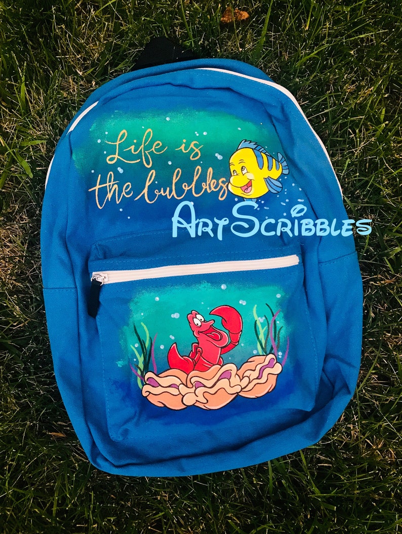 Pu&ograve; includere: Uno zaino in tela blu con un design dipinto a mano con i personaggi Sebastian e Flounder del film Disney La Sirenetta. Lo zaino ha il testo "Life is the bubbles" e "Art Scribbles" dipinto su di esso.