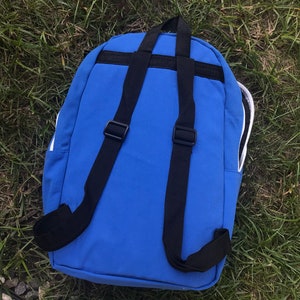 Pu&ograve; includere: Uno zaino blu con spalline nere e rifiniture bianche. Lo zaino ha una chiusura con cerniera e una piccola tasca sul davanti.