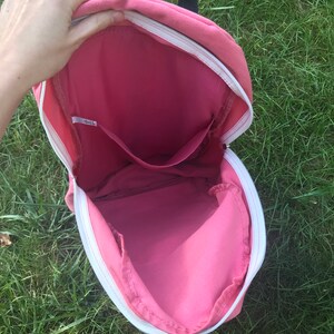 May include: Pink canvas backpack with a white zipper and a small pocket inside. The backpack is open and the inside is visible.