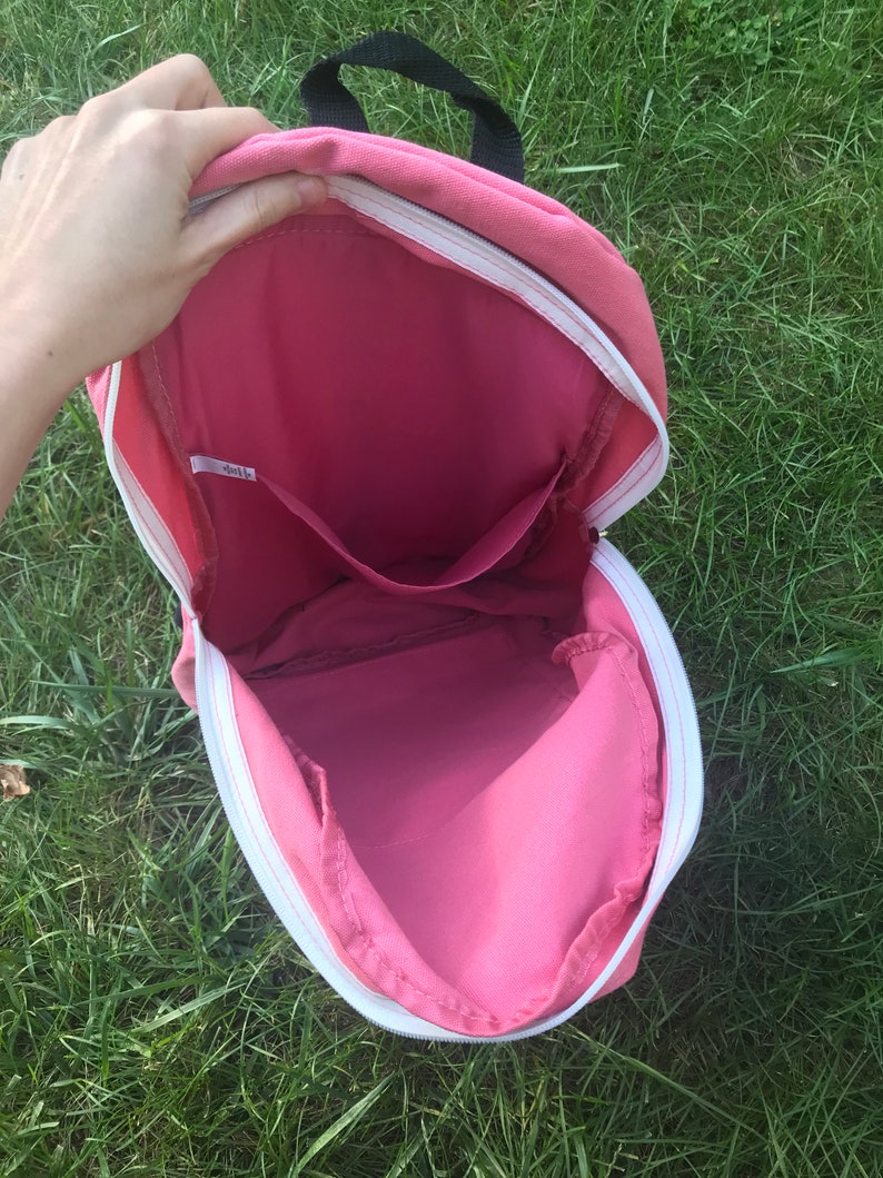 May include: Pink canvas backpack with a white zipper and a small pocket inside. The backpack is open and the inside is visible.