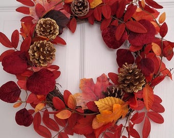 May include: A fall-themed wreath featuring a variety of red, orange, and yellow leaves. The wreath is adorned with several gold-painted pinecones. The leaves are arranged around a circular base, creating a festive and seasonal decoration.