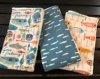 May include: Three fabric panels with different fishing-themed prints. The first panel has a white background with colorful fish, fishing lures, and the text "Gone fishing". The second panel has a blue background with white fish. The third panel has a white background with colorful fish lures and hooks.