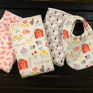 May include: Three baby bibs with farm animal prints. One bib has a pink pig pattern, one has a white sheep pattern, and one has a white background with a red barn, pigs, sheep, and chickens.