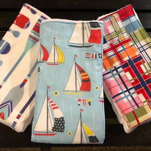 Op de afbeelding: Drie stoffen baby slabbetjes met nautische ontwerpen. Eén doek heeft blauwe en witte roeispanen, een andere heeft zeilboten op een lichtblauwe achtergrond en de derde heeft een patchwork ruitpatroon in rood, roze, oranje en blauw.