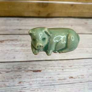 Może przedstawiać: Mała, jasnozielona ceramiczna figurka świnki. Świnka leży na boku, głowa odwrócona w bok. Świnka ma białą plamkę na boku.