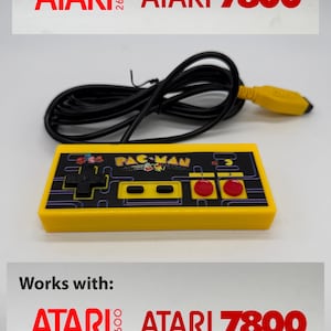 May include: Yellow Pac-Man video game controller with a black directional pad and red action buttons. The controller has a black cable and a yellow connector. The text "PAC-MAN" is visible on the controller. The text "ATARI 2600" and "ATARI 7800" are also visible.