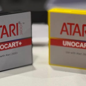 May include: Two Atari 2600+ Unocart+ cartridges. One is black, the other is yellow. Both have a gray label with the Atari logo in red, the text "UNOCART+", and "Use with Atari 2600+".