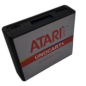 May include: Black Atari 2600+ UNOCART+ cartridge. The cartridge has a gray label with the Atari logo in red, the text "UNOCART+", and a red and yellow stripe. The text "Use with Atari 2600+" is also visible.