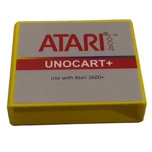 May include: A yellow Atari Unocart+ cartridge for the Atari 2600+ video game console. The cartridge has a gray label with the Atari logo in red, the text "UNOCART+", and the text "Use with Atari 2600+".