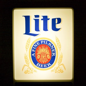 May include: A rectangular illuminated sign with the word "Lite" in blue, above a circular emblem that reads "A Fine Pilsner Beer." The sign has a cream-colored background and a digital clock displaying "1:00 AM" at the bottom.