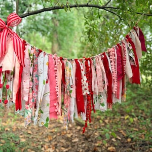 May include: A decorative fabric rag garland featuring a variety of hanging strips in shades of red, pink, and white. The garland includes floral patterns, gingham checks, and solid textures, finished with a large red and white striped bow on one end.