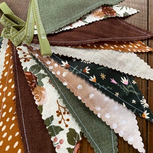 May include: A bunting banner with a variety of fabric pennants in shades of brown, green, and cream. The pennants feature different patterns, including floral prints, polka dots, and solid colors.