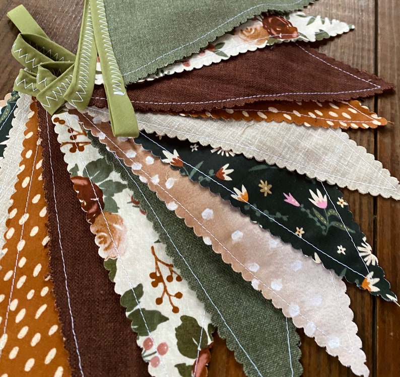 May include: A bunting banner with a variety of fabric pennants in shades of brown, green, and cream. The pennants feature different patterns, including floral prints, polka dots, and solid colors.