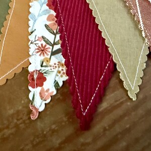 May include: A close-up of a decorative banner with various fabric pennants. The pennants feature a scalloped edge and are in shades of orange, green, red, and tan. Some pennants have floral patterns, while others are solid colors.