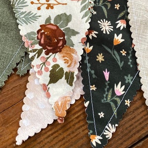 May include: A set of four fabric pennants with scalloped edges. The pennants are made of different fabrics with floral patterns in shades of green, brown, white, and pink. The pennants are sewn together to create a garland.