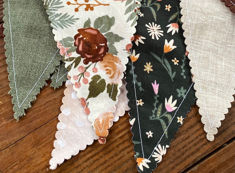 May include: A set of four fabric pennants with scalloped edges. The pennants are made of different fabrics with floral patterns in shades of green, brown, white, and pink. The pennants are sewn together to create a garland.