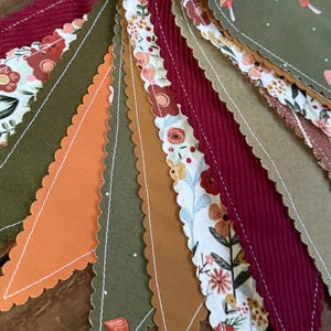May include: A colorful fabric banner with alternating triangular pennants. The pennants feature various patterns, including floral prints, corduroy, and solid colors in shades of orange, burgundy, olive green, and brown. A light yellow ribbon is at the top.