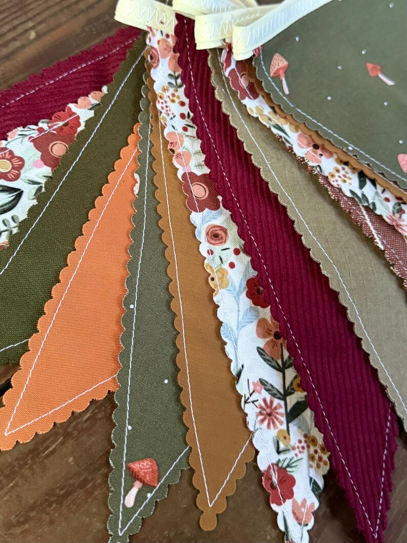 May include: A colorful fabric banner with alternating triangular pennants. The pennants feature various patterns, including floral prints, corduroy, and solid colors in shades of orange, burgundy, olive green, and brown. A light yellow ribbon is at the top.