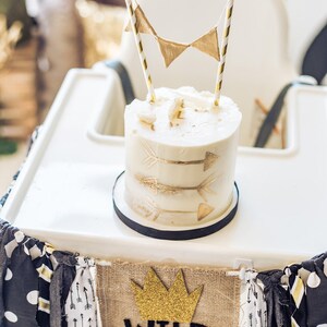 May include: A white cake with gold arrow designs and a gold crown banner that says "Wild One" on a white high chair.