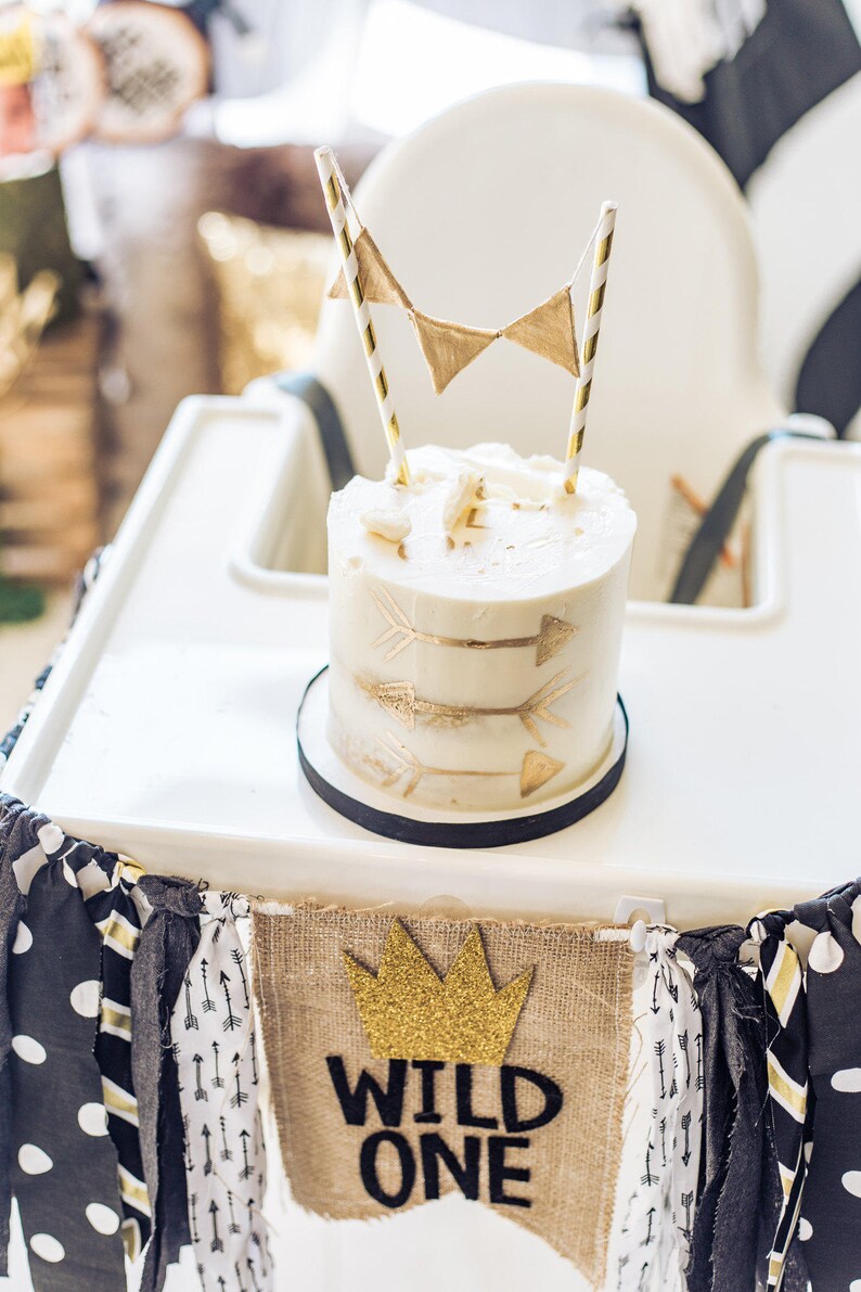 May include: A white cake with gold arrow designs and a gold crown banner that says "Wild One" on a white high chair.