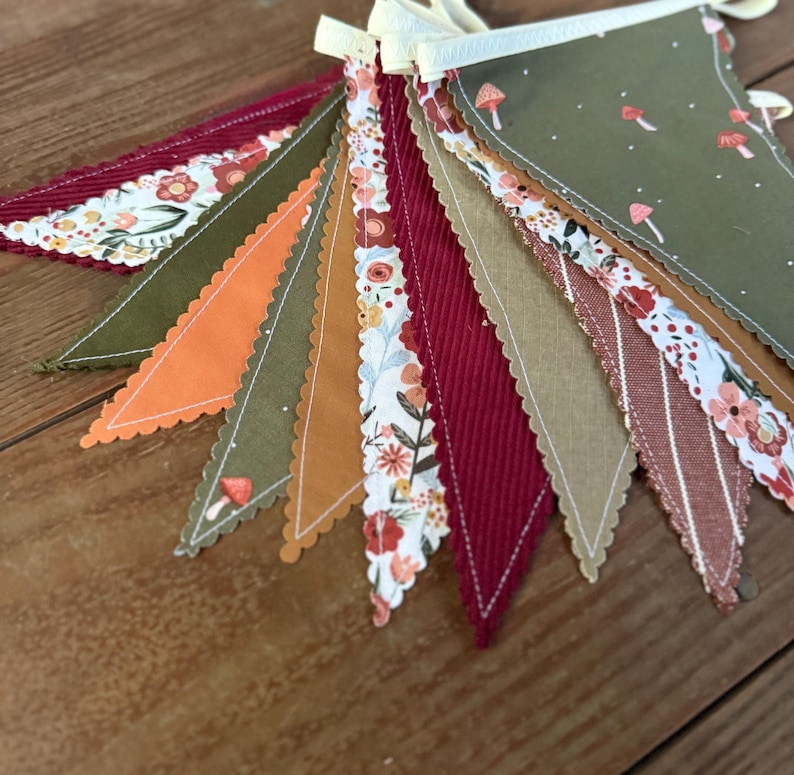 May include: A colorful fabric banner with various patterned pennants. The pennants feature floral, mushroom, and solid designs in shades of red, orange, green, and tan. The banner is strung on a cream-colored ribbon, perfect for festive decor.