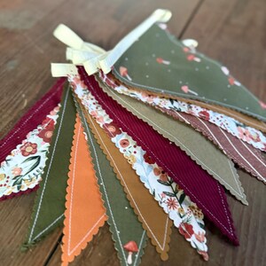May include: A colorful fabric banner with various patterned pennants. The pennants feature floral, mushroom, and striped designs in shades of green, orange, burgundy, and brown. The banner is tied with a yellow ribbon.