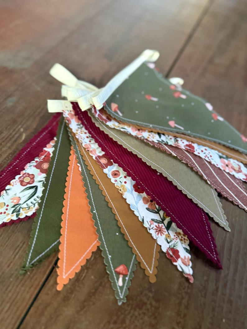 May include: A colorful fabric banner with various patterned pennants. The pennants feature floral, mushroom, and striped designs in shades of green, orange, burgundy, and brown. The banner is tied with a yellow ribbon.