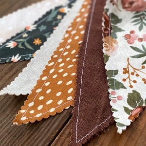 May include: A garland of fabric pennants with different patterns, including floral, polka dot, and solid colors. The pennants are sewn together with white thread and have a scalloped edge.