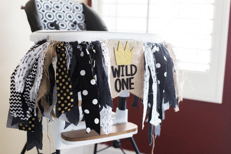 May include: A high chair decorated with a black, white and gold fabric banner that says "Wild One" with a gold glitter crown. The banner is attached to the high chair with a hessian ribbon.