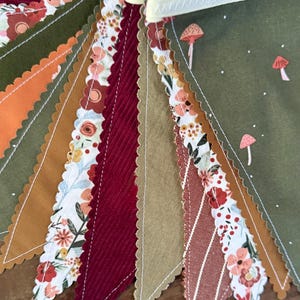 May include: A colorful fabric bunting with various patterns and textures. The pennants feature floral, mushroom, and striped designs in shades of green, orange, burgundy, and tan. The bunting is strung on a cream-colored ribbon.