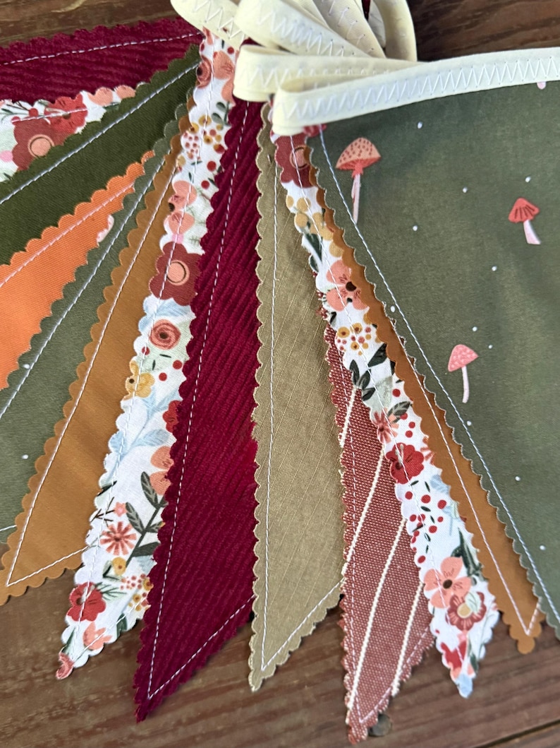 May include: A colorful fabric bunting with various patterns and textures. The pennants feature floral, mushroom, and striped designs in shades of green, orange, burgundy, and tan. The bunting is strung on a cream-colored ribbon.