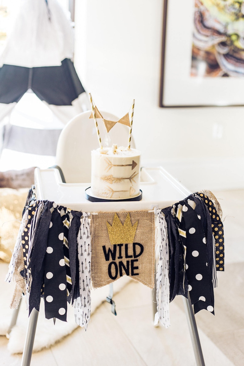 May include: A white cake with gold arrows and a gold crown on top, decorated with a hessian banner that says "Wild One". The cake is on a white high chair with a black and white fabric garland.