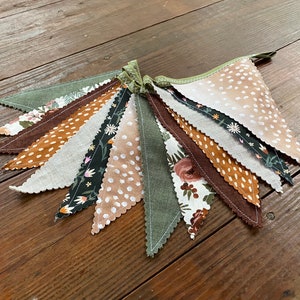 May include: A bunting banner with 10 flags in various patterns. The flags are made of fabric and feature a variety of colors, including brown, green, white, and black. The flags are sewn together with a green thread.