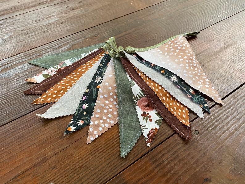 May include: A bunting banner with 10 flags in various patterns. The flags are made of fabric and feature a variety of colors, including brown, green, white, and black. The flags are sewn together with a green thread.